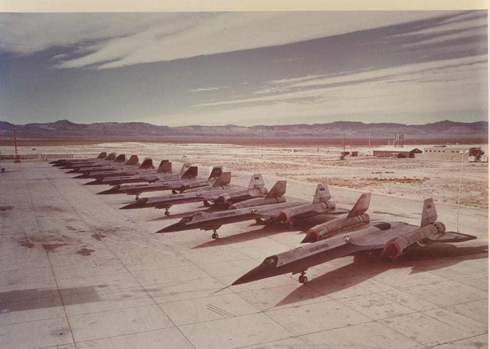 A row of A-12 aircraft parked on a desert ramp, unpainted titanium with dark leading edges.