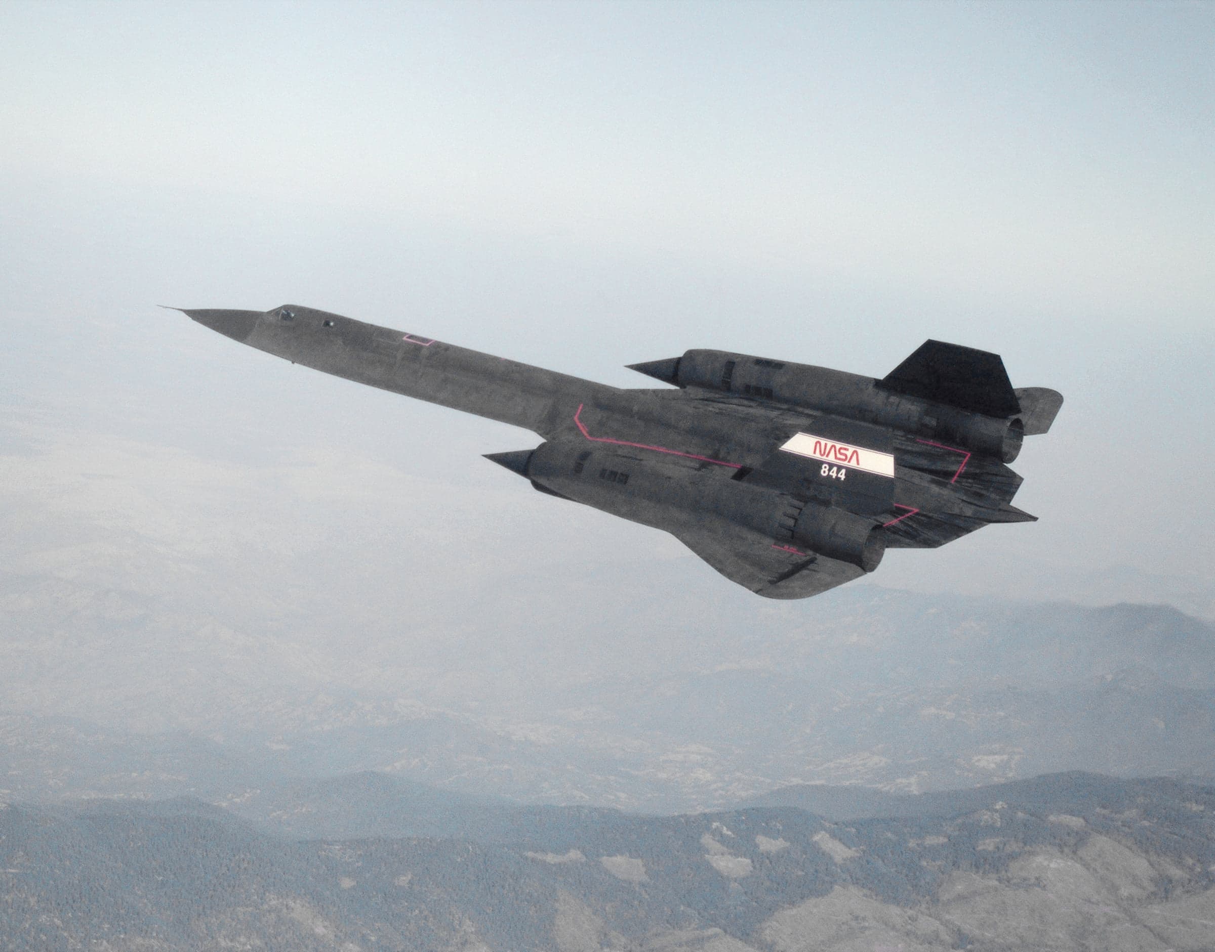 NASA SR-71A tail number 844 in flight over mountains, with two cockpit canopies in tandem.
